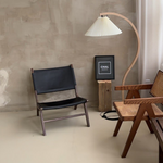 Load image into Gallery viewer, Contemporary interior design featuring the Cozy Glow Lamp with an arched wooden frame and pleated shade, alongside a collection of eclectic wooden chairs against a textured concrete wall, creating a modern, artistic ambiance.
