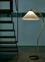 Load image into Gallery viewer, The Cozy Glow Lamp illuminates a minimalist setting beside a modern staircase, featuring its iconic curved wooden frame and pleated lampshade, creating a sophisticated and artistic lighting element in a contemporary space.
