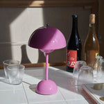 Load image into Gallery viewer, A vibrant pink rechargeable LED desk lamp illuminating a space with a playful touch. The lamp is surrounded by two wine bottles, two textured glasses, and a neatly placed book with a golden design. Sunlight casts a warm glow on the items, creating a cozy and inviting atmosphere.
