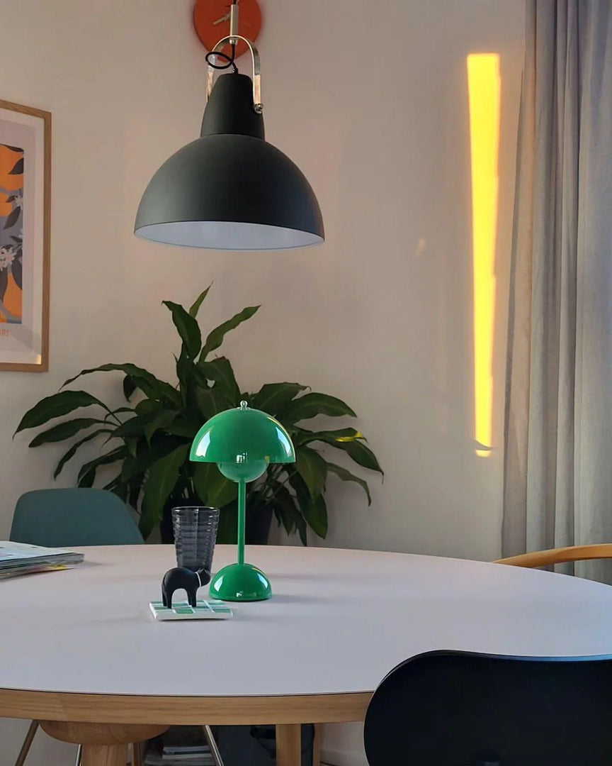 A vibrant green rechargeable LED small desk lamp placed on a minimalist white table, accompanied by a glass, a small black elephant sculpture, and a stack of books. In the background, a plant adds a natural touch, while a hanging black pendant light illuminates the space. The soft sunlight streaming through the window enhances the cozy ambiance.