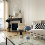 Load image into Gallery viewer, A stylish blue rechargeable LED small desk lamp is placed on a glass coffee table in a cozy living room. The room features a beige sofa with striped cushions, a decorative plant, and a candle. A fireplace and a mirror adorn the wall, creating a welcoming atmosphere.
