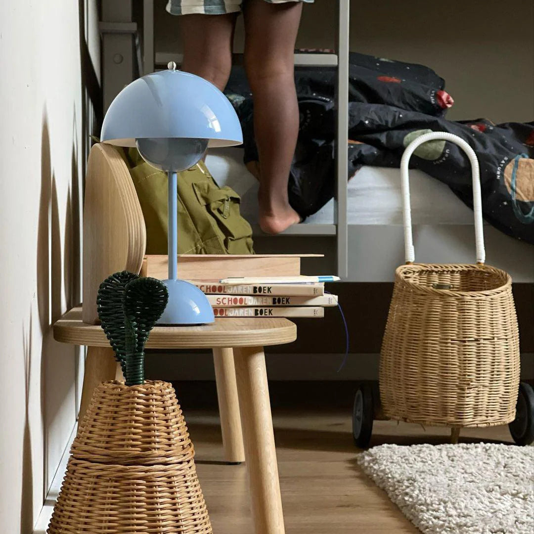 A blue rechargeable LED small desk lamp rests on a wooden table, next to a stack of books titled "School" in Dutch. The lamp stands beside a woven basket, and a child’s legs can be seen standing near a bed in the background. The room has a cozy, personal vibe, with soft natural light illuminating the items in the foreground.