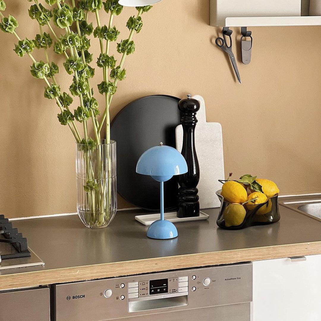 A blue rechargeable LED small desk lamp is placed on a kitchen counter next to a vase of fresh flowers, a black plate, and a bowl of lemons. The kitchen features a Bosch dishwasher and sleek countertops, with a modern and minimalistic aesthetic accentuated by the warm tones of the wall and decorative items.