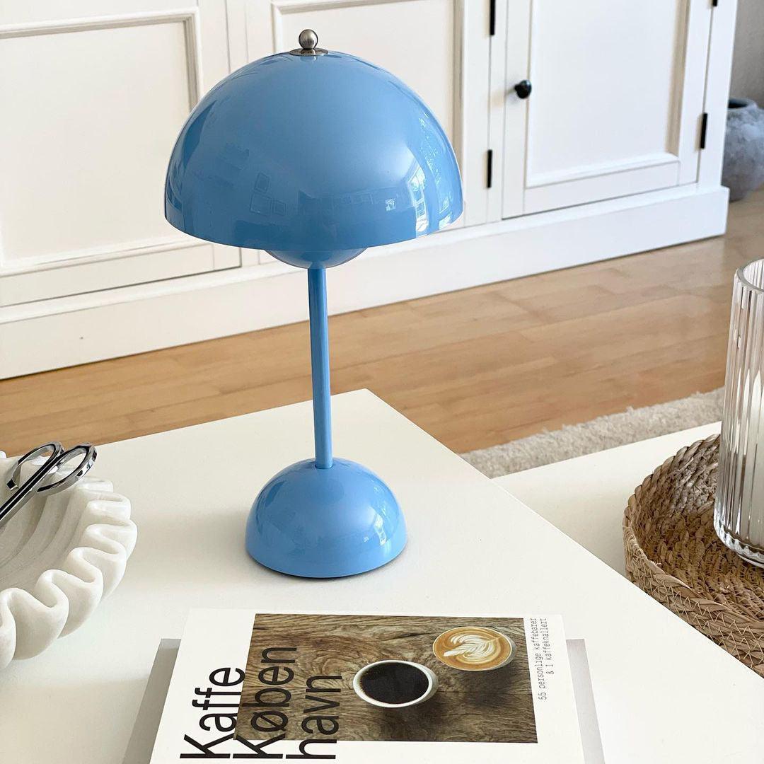 A blue rechargeable LED small desk lamp is placed on a coffee table next to a book titled "Kaffe Koebenhavn" and a woven basket. The lamp features a modern and minimalist design, providing a playful yet sophisticated look to the space with the light wooden floor and simple white surroundings.