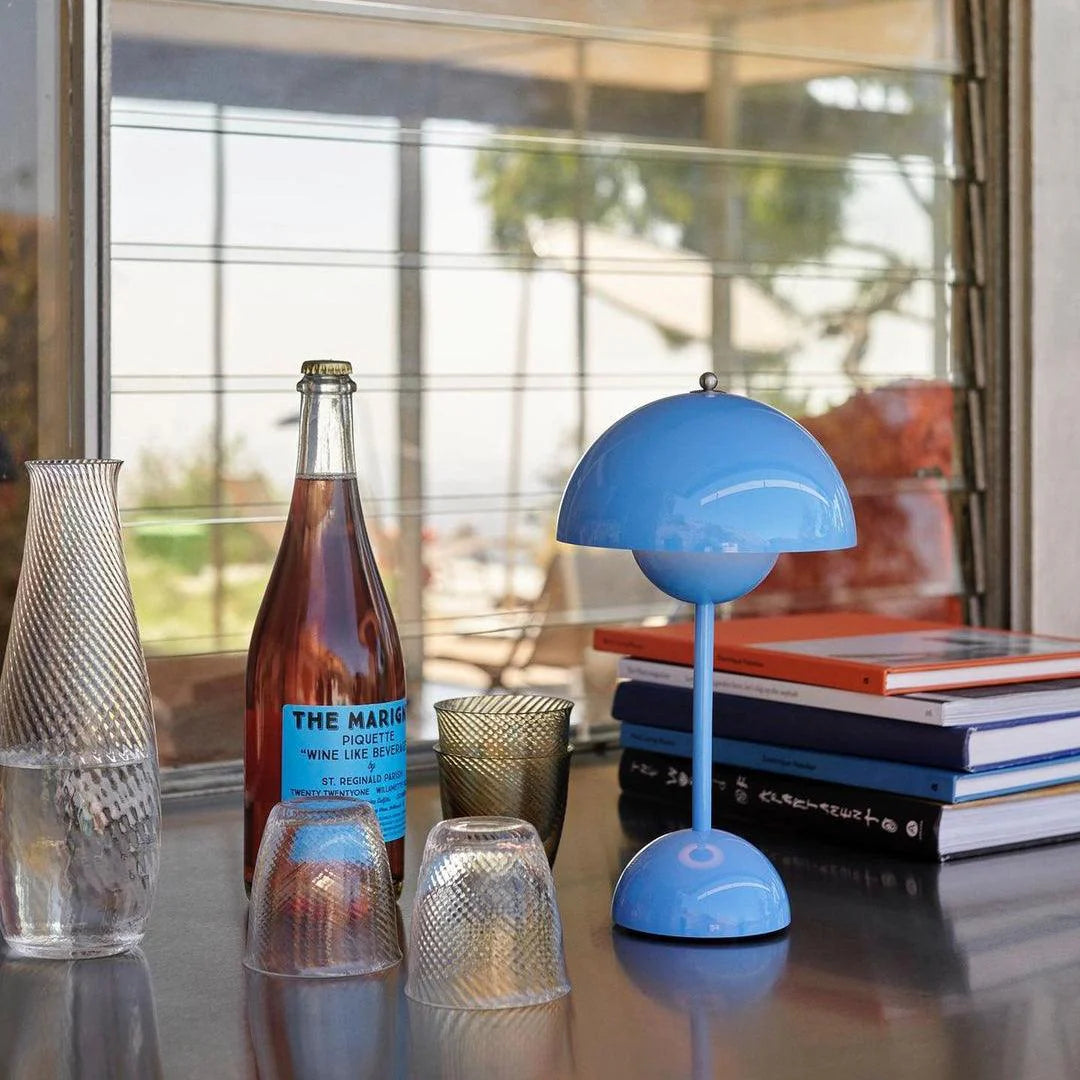 A blue rechargeable LED small desk lamp stands on a table beside a bottle of wine labeled "The Marigny," a few glasses, and several stacked books. The table is surrounded by a bright and open outdoor setting, with palm trees visible through the window in the background. The lamp's sleek design adds a touch of contemporary style to the vibrant atmosphere.