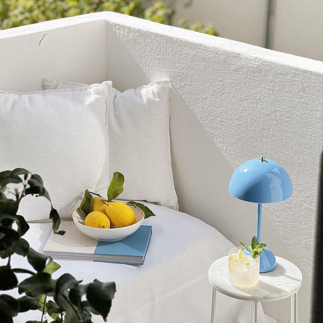 A vibrant blue rechargeable LED small desk lamp placed on a marble table beside a refreshing drink with mint leaves, accompanied by a bowl of lemons and a couple of blue books. The setting exudes a serene and sunny atmosphere with white pillows and soft lighting, creating a peaceful outdoor vibe.