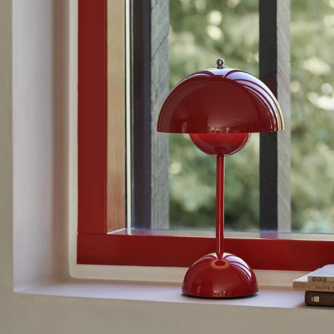 A red rechargeable LED small desk lamp sits elegantly on a windowsill, with a lush green view outside. The vibrant red lamp contrasts with the window frame and surrounding greenery, creating a bold and eye-catching aesthetic.