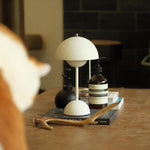 Load image into Gallery viewer, A white rechargeable LED small desk lamp is placed on a wooden table along with Aesop skincare bottles, stacked books, and a wooden branch. The warm, inviting scene includes a partially visible cat in the foreground, creating a calm, homely atmosphere.
