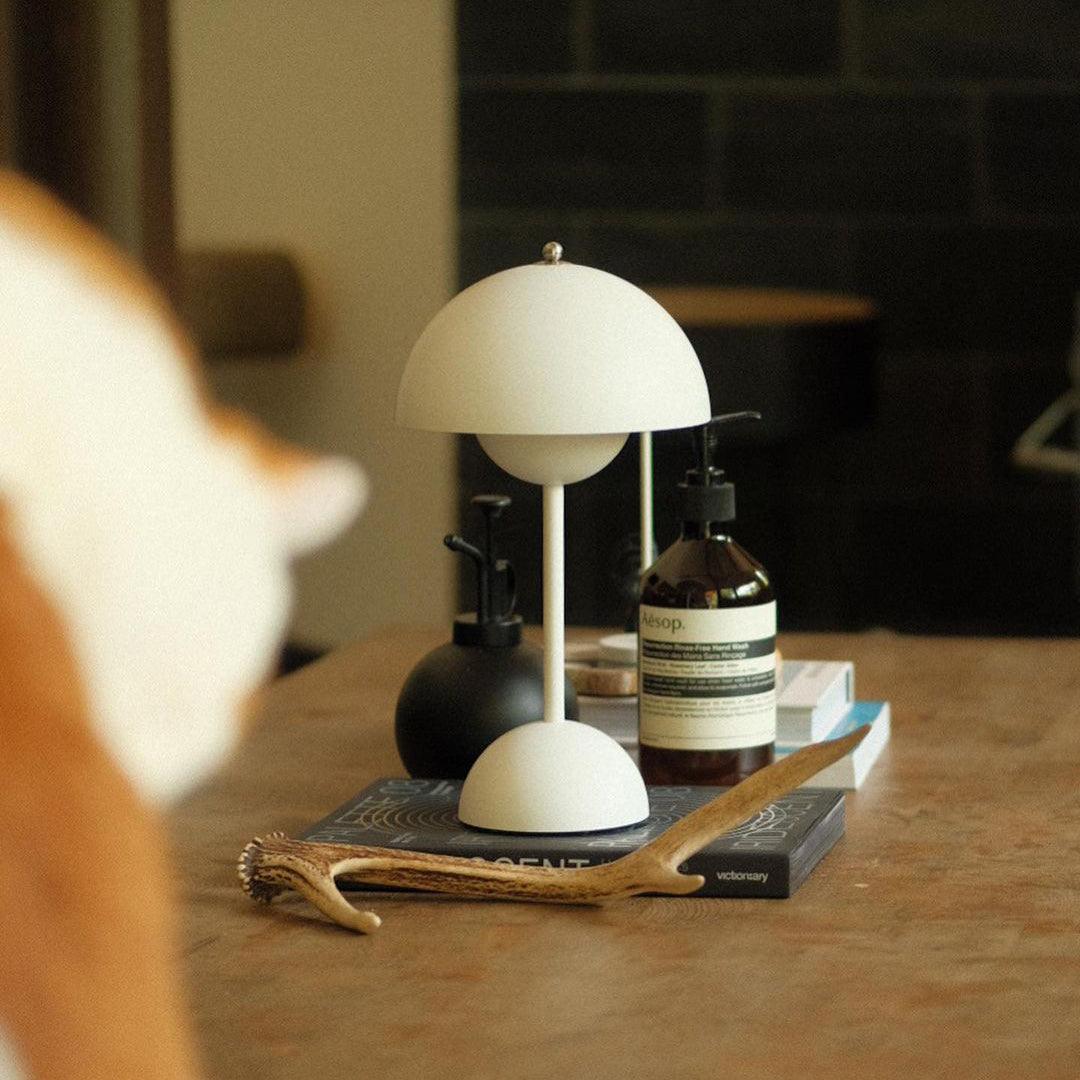 A white rechargeable LED small desk lamp is placed on a wooden table along with Aesop skincare bottles, stacked books, and a wooden branch. The warm, inviting scene includes a partially visible cat in the foreground, creating a calm, homely atmosphere.