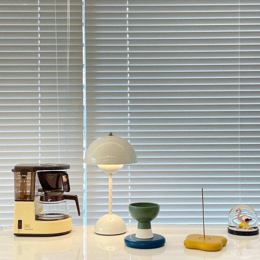 A white rechargeable LED small desk lamp is placed on a clean countertop, surrounded by a coffee maker, a colorful incense holder, a decorative glass dome, and a green and blue sculpture, all against a backdrop of blinds.