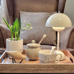 Load image into Gallery viewer, A white rechargeable LED small desk lamp sits on a wooden tray surrounded by a ceramic jar, a small bird figurine, a plant in a pot, and a coffee cup with a spoon. The scene is cozy, evoking a sense of warmth and calm, with a comfortable armchair in the background.
