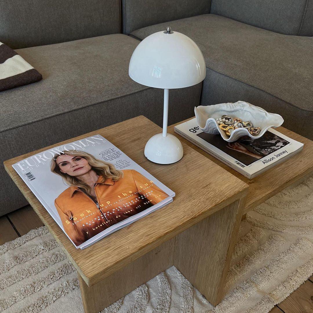 A white rechargeable LED small desk lamp rests on a wooden coffee table, next to fashion magazines and a decorative dish. The lamp’s sleek, minimal design complements the neutral tones of the living room, with the couch and surrounding decor adding a cozy atmosphere.