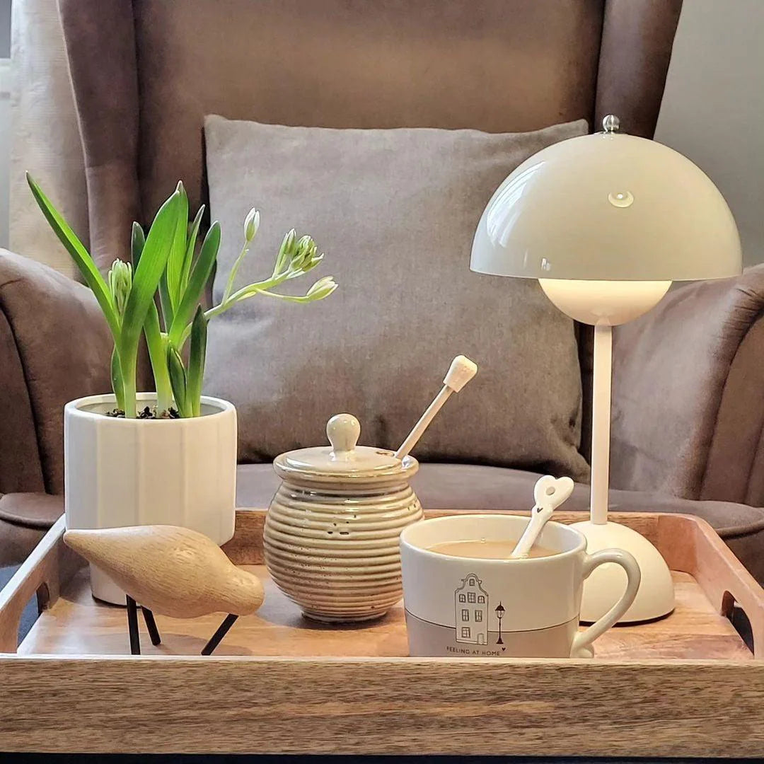 A white rechargeable LED small desk lamp sits on a wooden tray surrounded by a ceramic jar, a small bird figurine, a plant in a pot, and a coffee cup with a spoon. The scene is cozy, evoking a sense of warmth and calm, with a comfortable armchair in the background.