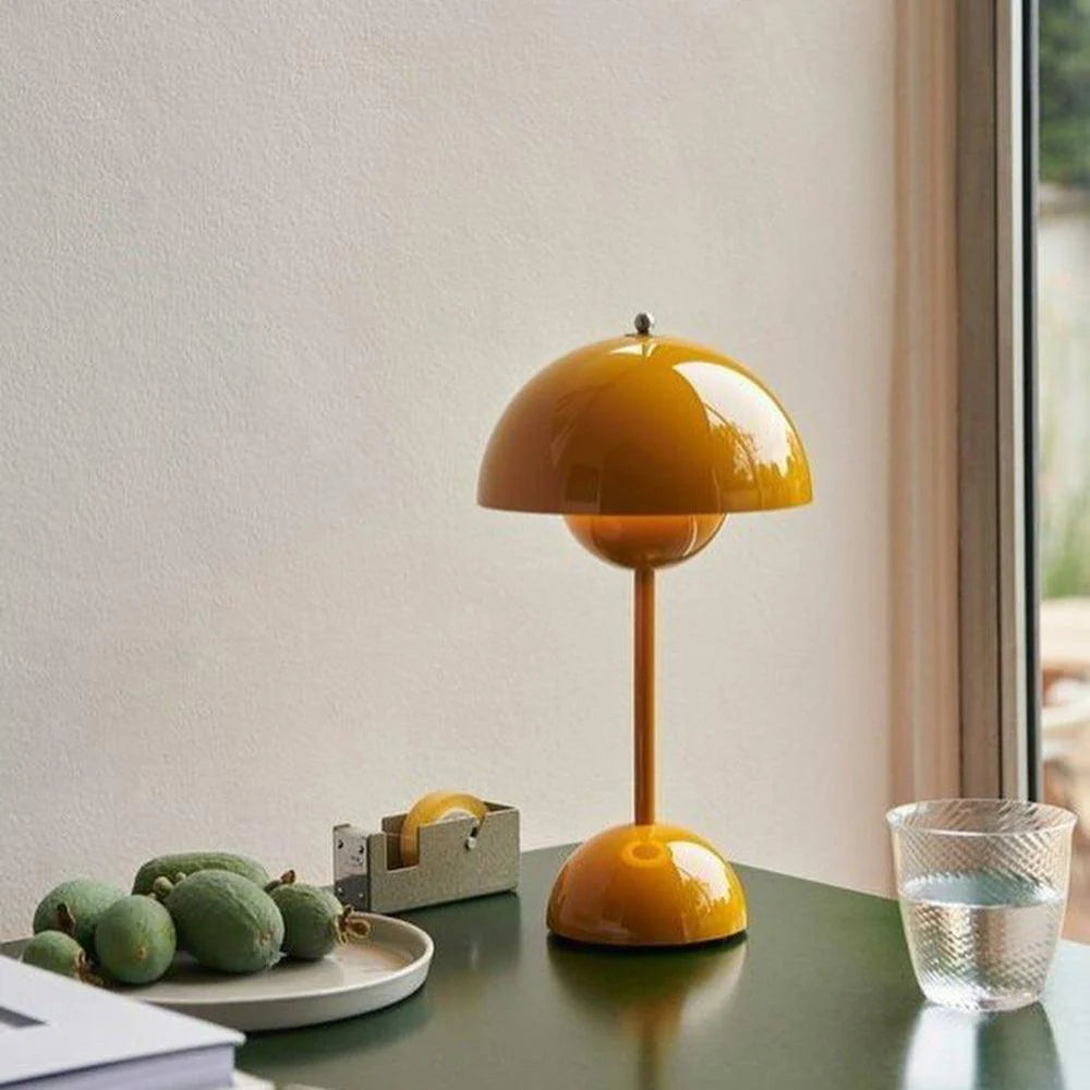 A vibrant yellow rechargeable LED small desk lamp rests on a desk next to a plate with green kiwifruits and a tape dispenser. The lamp’s dome-shaped shade and base match in glossy yellow, creating a cheerful ambiance. Nearby, a glass of water sits next to the window, offering a glimpse of natural light and outdoor greenery.