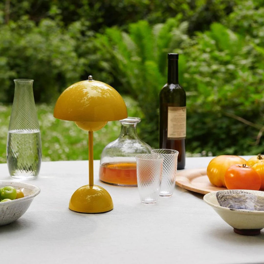 A rechargeable LED small desk lamp in bright yellow sits on a rustic table set outdoors. The lamp’s dome shade and rounded base reflect the sunlight as it complements a laid-back gathering with fruits, a wine bottle, and crystal glasses. Lush green foliage can be seen in the background, enhancing the natural setting.