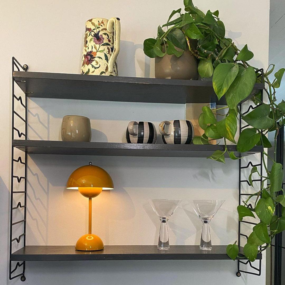 A rechargeable LED small desk lamp in yellow illuminates a stylish shelf. The lamp rests beside a collection of ceramics and clear glassware, with a vibrant green plant cascading from the top shelf. The light casts a warm glow in the cozy and artistic setting.