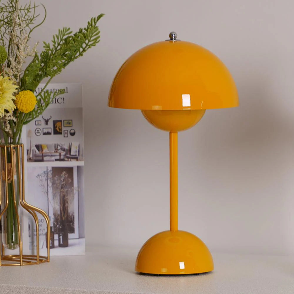 A bright yellow rechargeable LED small desk lamp stands on a light-colored surface. The lamp features a round, shiny yellow dome and base with a slim stem. Nearby, a vase of fresh flowers, including yellow blooms, adds a natural touch to the modern setup. A framed photo or illustration is partially visible in the background.