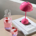 Load image into Gallery viewer, A stylish pink rechargeable LED lamp beside a glass and a carafe filled with a refreshing pink drink, ice cubes, and raspberries. The items are placed on a white book with a chic design, and the warm sunlight highlights the vibrant colors, giving a lively and inviting vibe to the scene.
