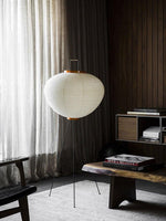 Load image into Gallery viewer, This floor lamp features a beautiful round rice paper shade with a pleated design, perched on black tripod legs. The lamp’s simple, minimalist appearance blends seamlessly into a cozy room with rich wood paneling and soft curtains, providing a calm and warm ambiance.
