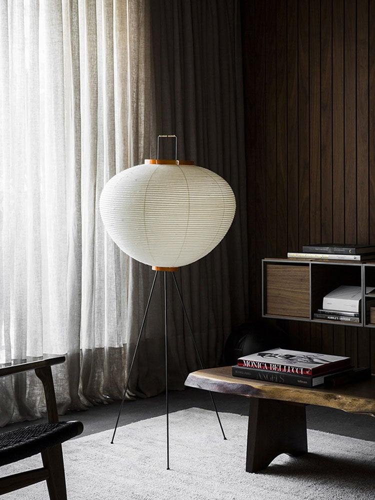 This floor lamp features a beautiful round rice paper shade with a pleated design, perched on black tripod legs. The lamp’s simple, minimalist appearance blends seamlessly into a cozy room with rich wood paneling and soft curtains, providing a calm and warm ambiance.