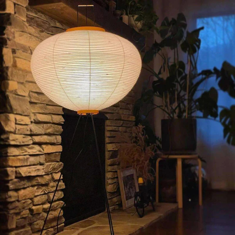 The rice paper floor lamp softly illuminates the stone fireplace in a cozy, plant-filled living space. Its minimalist, rounded design contrasts beautifully with the rustic stone wall and adds a serene atmosphere to the room. The lamp's gentle light enhances the relaxed ambiance as evening falls, creating a perfect setting for comfort and style.