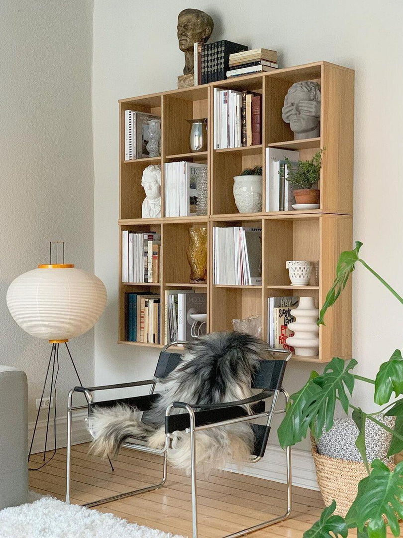 The rice paper floor lamp stands elegantly beside a modern wooden bookshelf filled with books and small sculptures. Its round, soft-lit shade creates a warm ambiance in the room, complementing the neutral-toned furnishings and plant. The minimalist design of the lamp enhances the cozy yet sophisticated aesthetic of the space.