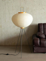 Load image into Gallery viewer, A stylish rice paper floor lamp stands on black tripod legs with a simple yet elegant design. The round, soft-lit lampshade is made from delicate, pleated rice paper and is accented with a wooden ring at the top and bottom. It illuminates a cozy, minimalist space with warm, diffused light.
