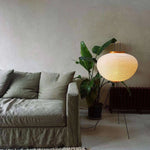 Load image into Gallery viewer, This rice paper floor lamp casts a soft, warm glow from its round, textured shade. Resting on a minimalist metal frame, it adds a cozy and serene feel to a room with its calm yellow lighting. The lamp sits next to a comfortable green sofa and a lush indoor plant, creating an inviting atmosphere.
