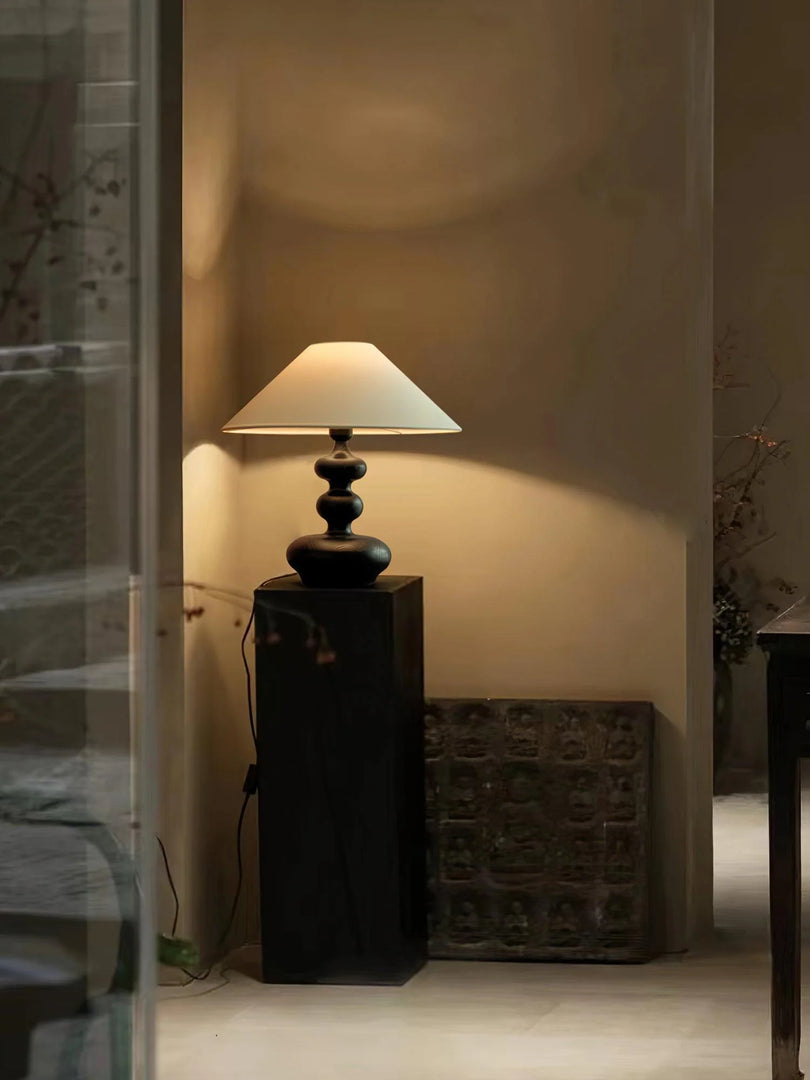 An evocative ambient corner illuminated by a Gourd Lamp with a stacked dark wooden base and a classic white shade. Positioned on a tall, slender pedestal, the lamp casts a soft, warm light against a textured wall, accompanied by an antique decorative panel nearby, creating an inviting and mysterious atmosphere in the room.