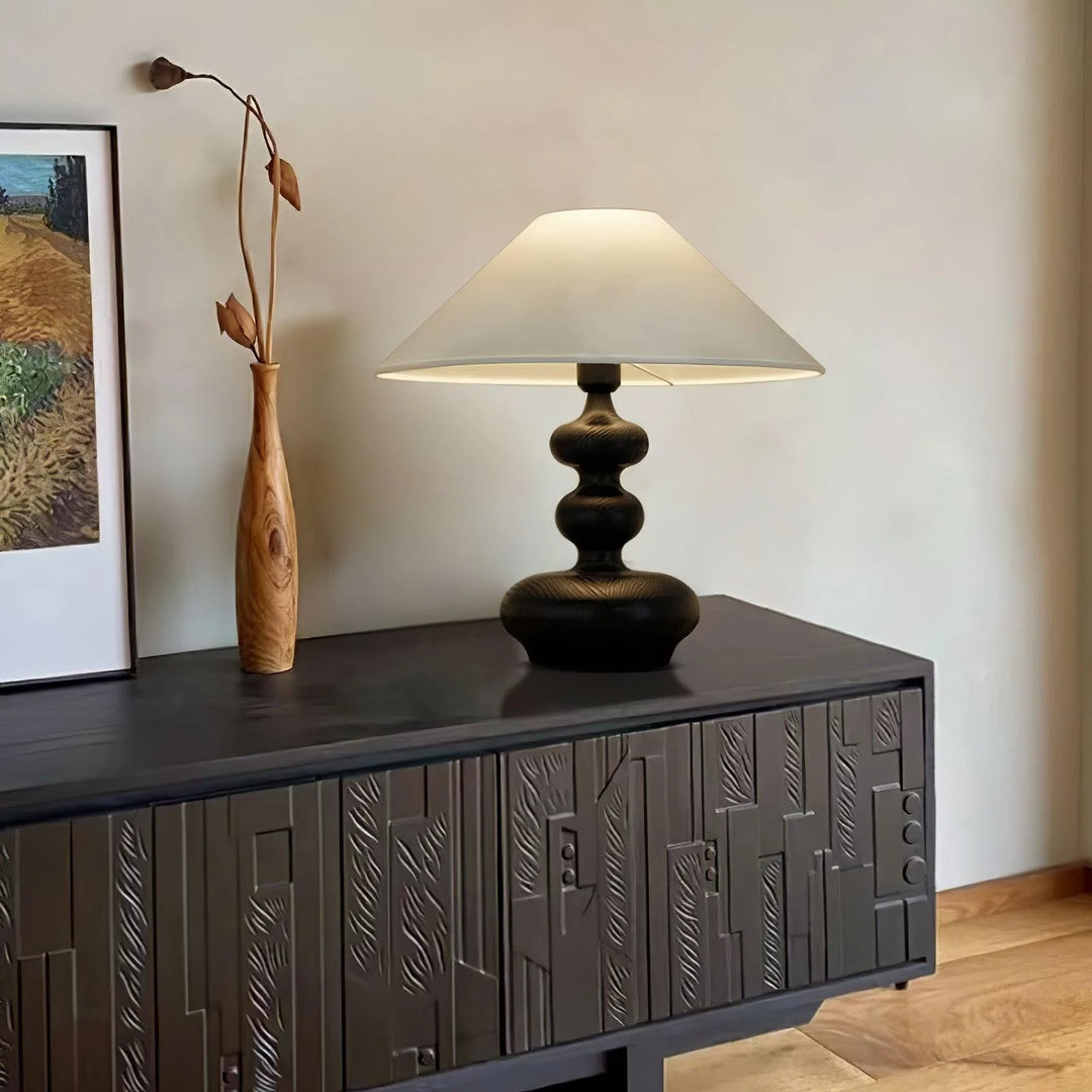 An artistic display featuring a Gourd Lamp with a dark wooden base and a simple white shade, elegantly placed on a textured black sideboard. Accompanied by a tall wooden sculpture and a framed landscape painting, the setup creates a harmonious blend of natural textures and soft lighting, enriching the calm and cultured aesthetic of the space.