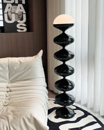 Load image into Gallery viewer, A stylish setting with an Artistic Gourd Lamp, featuring a towering black glossy base with smooth, spherical sections and a white dome light, set beside a plush white couch. A modern art piece with bold lettering hangs in the background, complementing the elegant, monochrome theme of the room.
