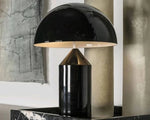 Load image into Gallery viewer, Atollo metal table lamp with a glossy black dome shade and a cylindrical base, elegantly displayed on a black marble surface with modern wall art in the background.
