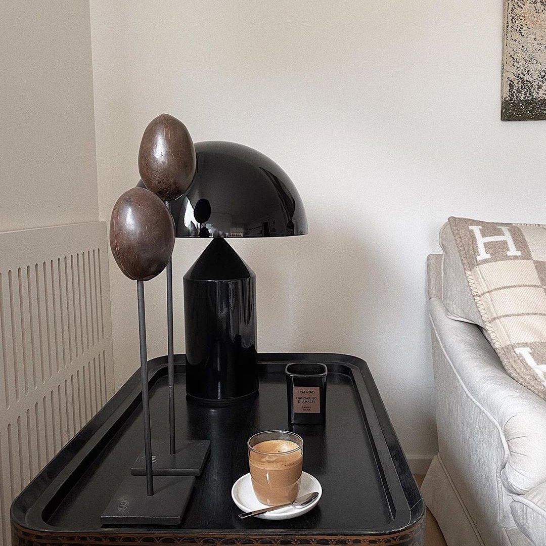 Atollo metal table lamp with a sleek black dome shade and cylindrical base styled on a black side table, accompanied by modern sculptures, a luxury candle, and a cup of coffee for an elegant and cozy living room setting.