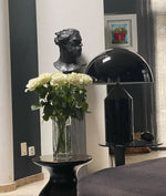 Load image into Gallery viewer, Atollo metal table lamp with a black dome shade and cylindrical base placed on a sleek side table, styled alongside a glass vase of white roses, with a black sculpture and framed artwork in the background for a sophisticated interior decor.
