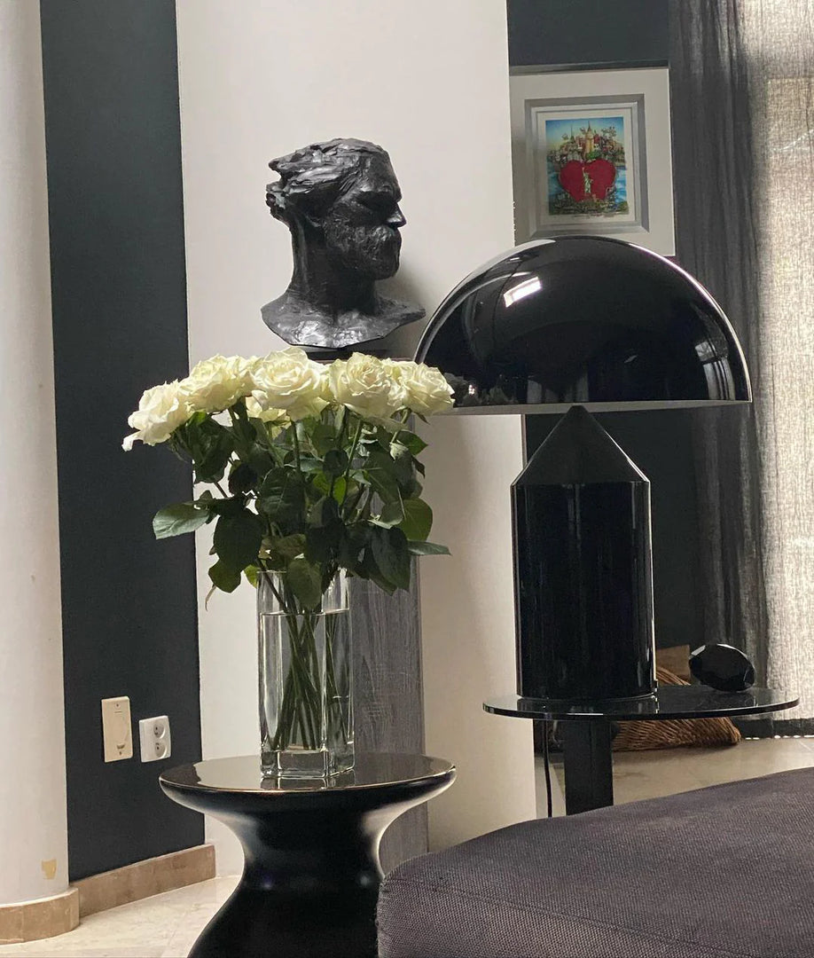 Atollo metal table lamp with a black dome shade and cylindrical base placed on a sleek side table, styled alongside a glass vase of white roses, with a black sculpture and framed artwork in the background for a sophisticated interior decor.