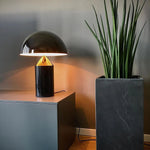 Load image into Gallery viewer, Atollo Metal Table Lamp in black, elegantly illuminating a space with its unique candle-shaped light bulb, placed next to a tall snake plant in a sleek marble pot, enhancing a modern office environment.
