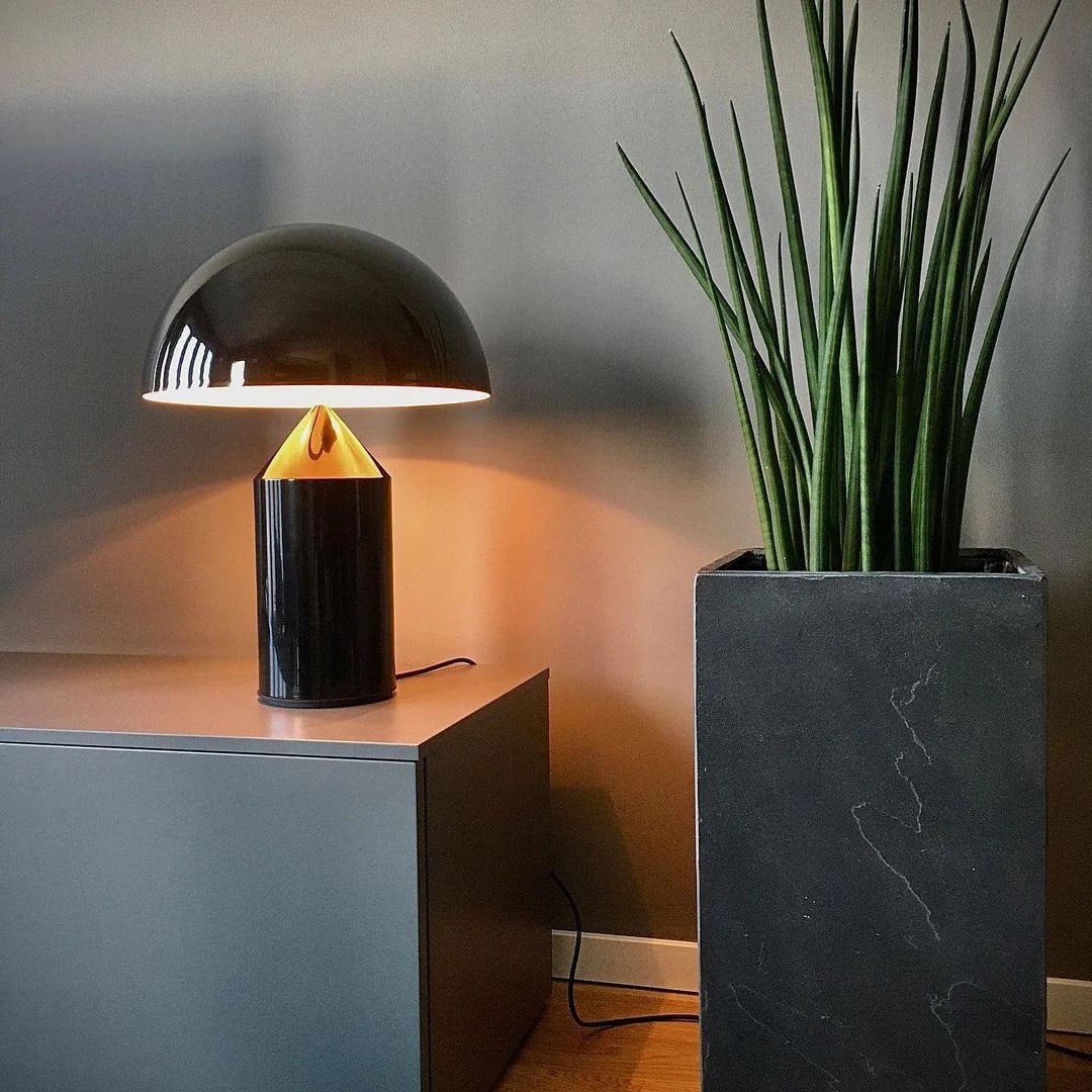 Atollo Metal Table Lamp in black, elegantly illuminating a space with its unique candle-shaped light bulb, placed next to a tall snake plant in a sleek marble pot, enhancing a modern office environment.