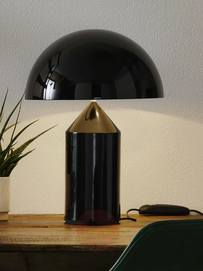 Atollo Metal Table Lamp in black on a wooden table, illuminated by natural light with a green plant in the background, adding a touch of nature to the modern and sleek design of the lamp.