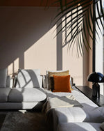 Load image into Gallery viewer, Atollo Metal Table Lamp in black, set on a side table beside a luxurious gray sofa with plush cushions under a sunlit window casting shadows of palm leaves, creating a tranquil and stylish living space.
