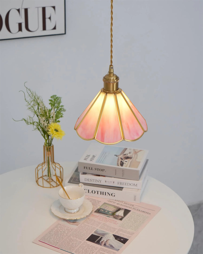 Butterfly pendant lamp with a glowing pink stained glass scalloped shade and gold accents, hanging above a white round table decorated with books, a teacup, a vase with yellow flowers, and a magazine, creating a stylish and cozy reading nook.