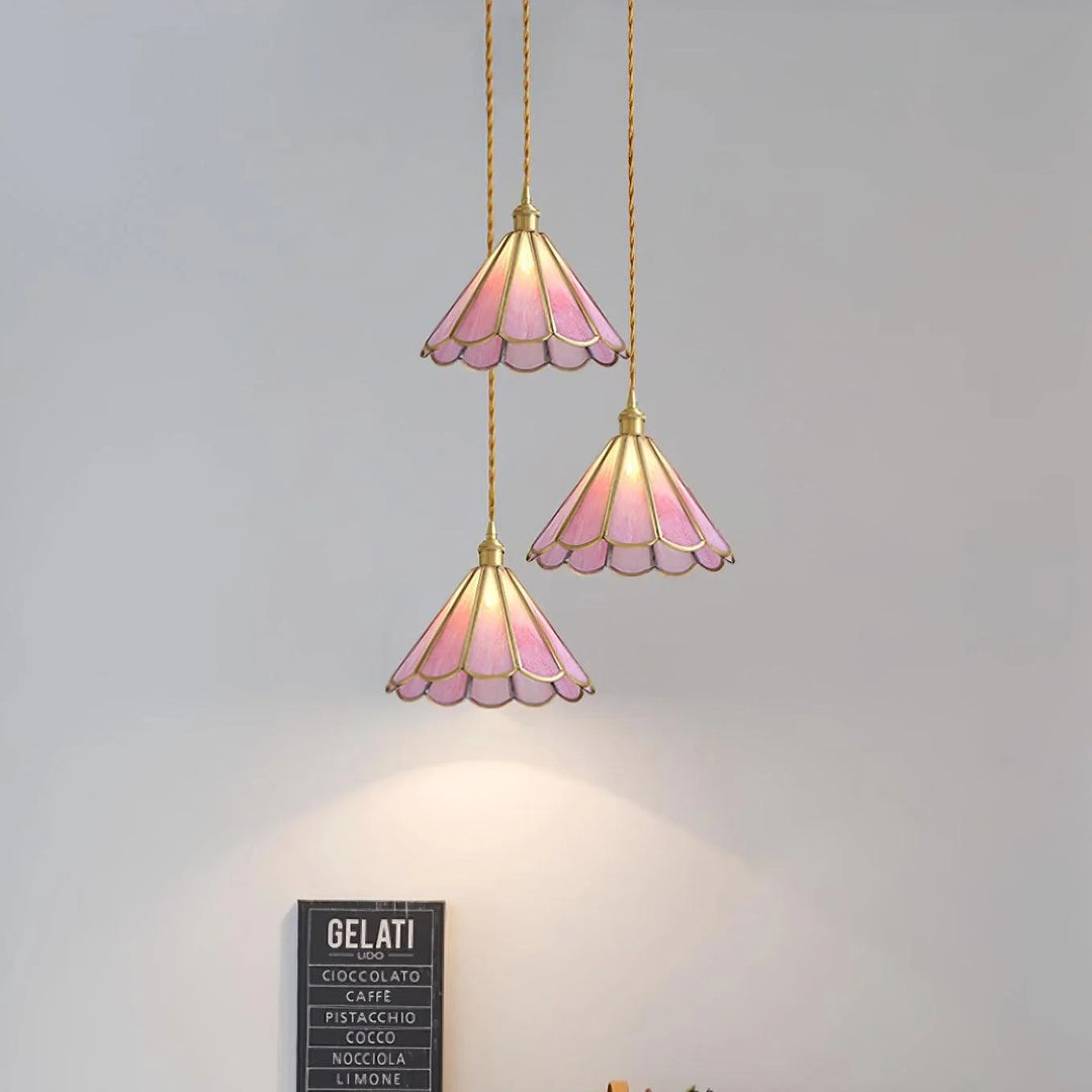 Trio of Butterfly pendant lamps with pink stained glass scalloped shades and gold accents, elegantly suspended at different heights on twisted cords, illuminating a wall with soft, warm light in a contemporary setting.