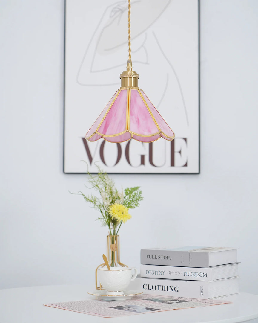 Butterfly pendant lamp with a pink stained glass scalloped shade and gold metal accents, hanging above a white table decorated with a teacup, books, and a vase of yellow flowers, with Vogue wall art adding a stylish backdrop.