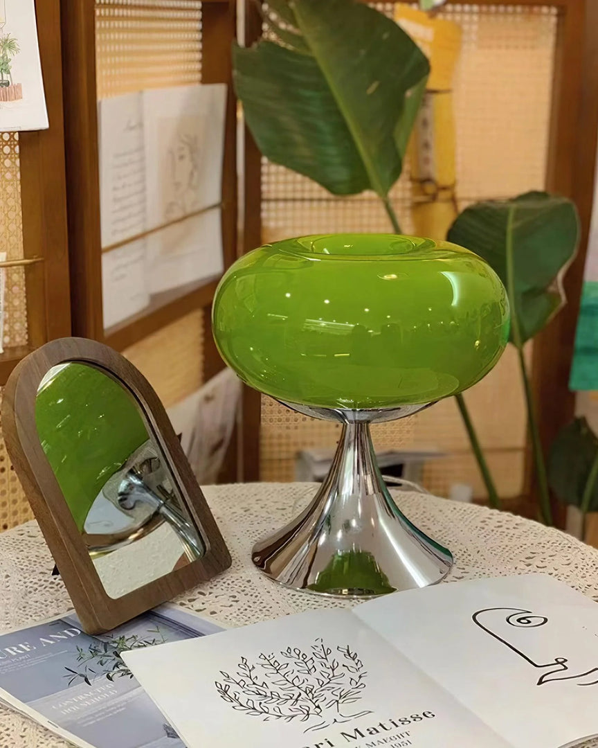 Candle table lamp with a vibrant green glass dome-shaped shade and polished chrome base, placed on a lace-covered table alongside an open art book and a wooden-framed mirror, with large green leaves in the background, creating an artistic and cozy decor scene.