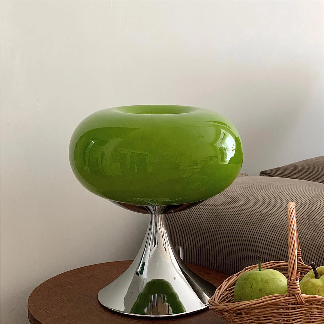 Candle table lamp with a glossy green glass dome-shaped shade and polished chrome base, placed on a wooden side table next to a wicker basket of green apples, adding a modern and fresh decor accent.