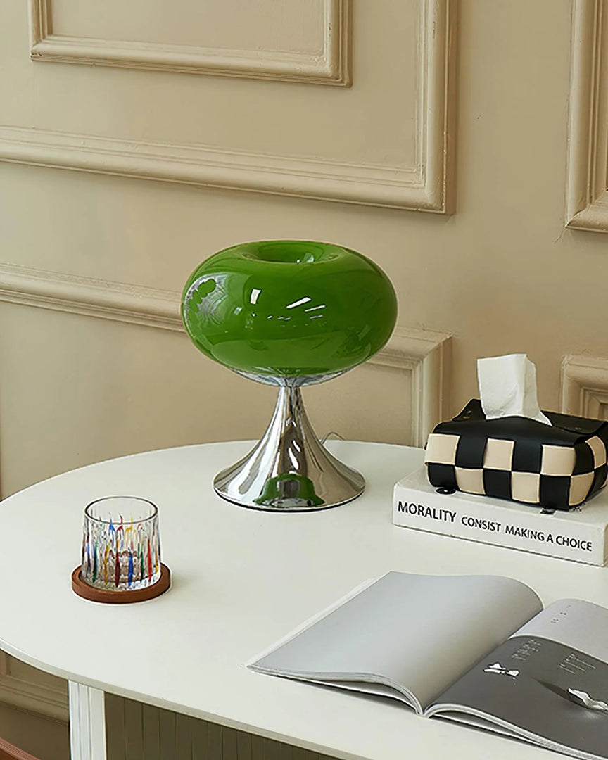 Candle table lamp with a glossy green glass dome-shaped shade and polished chrome base, styled on a white desk with an open magazine, a colorful glass candle holder, and a checkered tissue box, creating a modern and eclectic study space.