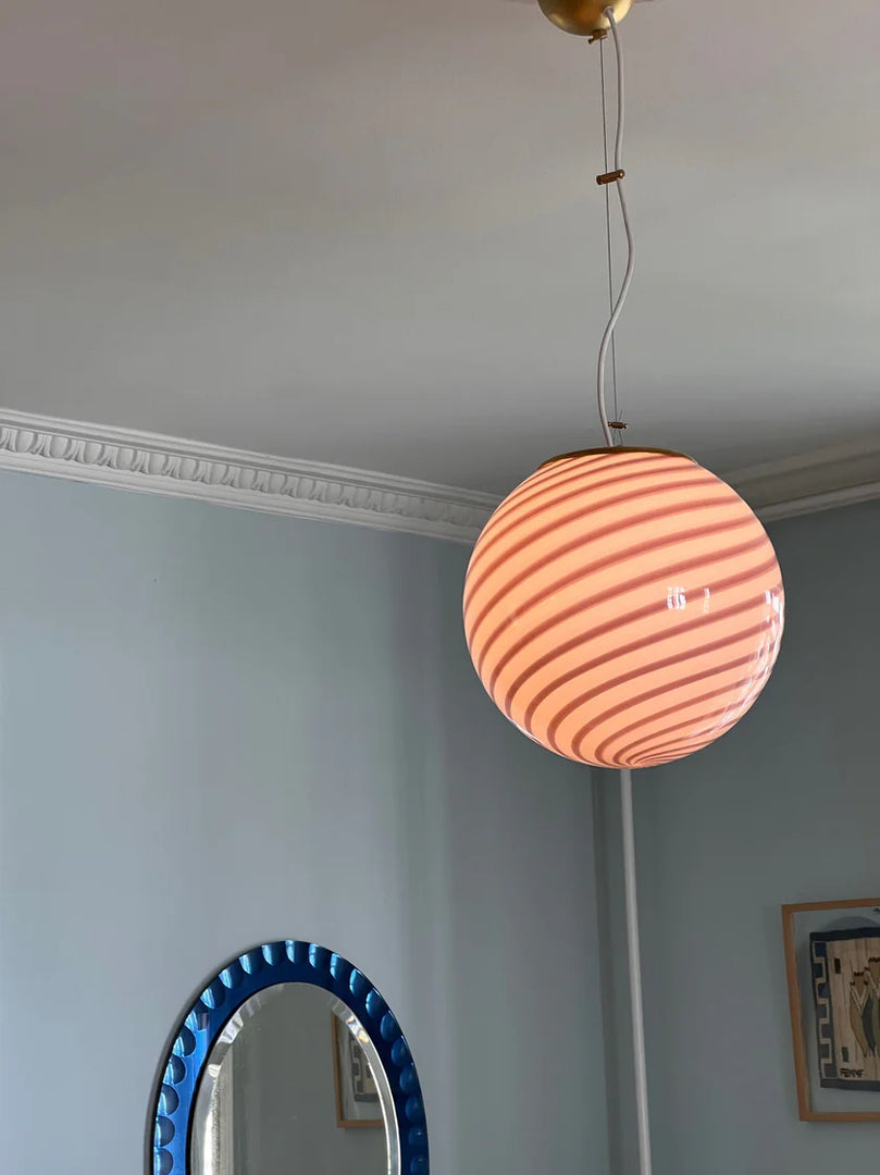 Candy pendant lamp with a soft pink and white striped globe-shaped shade, suspended from the ceiling, complemented by a blue circular mirror and light pastel walls, adding a whimsical touch to the room.