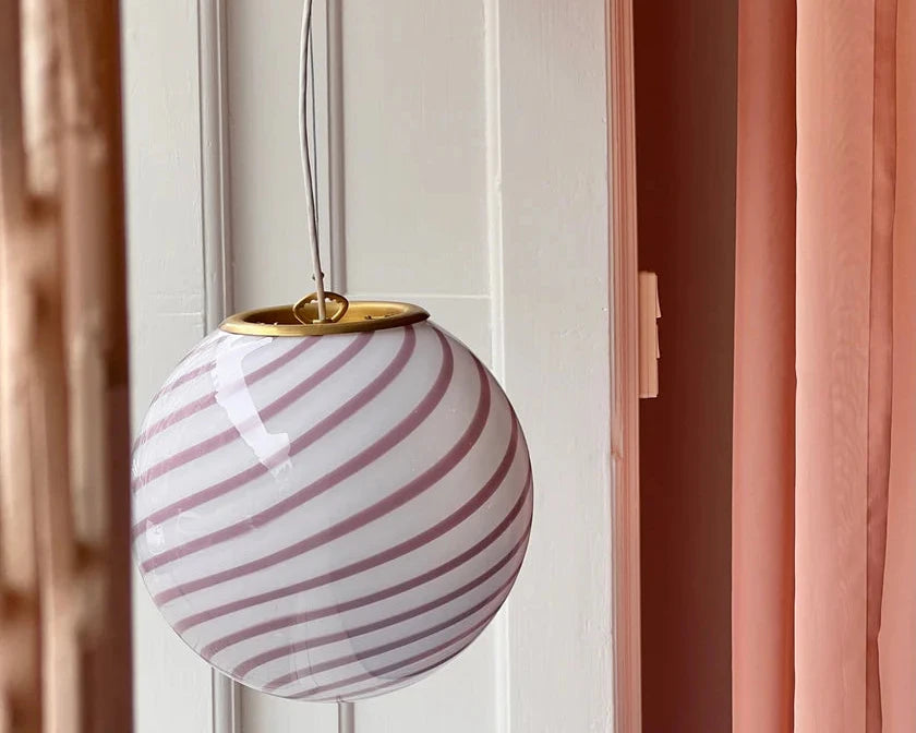 Candy pendant lamp featuring a soft pink and white striped globe-shaped glass shade with a sleek gold rim, suspended near soft pink curtains, adding a chic and contemporary touch to the room.