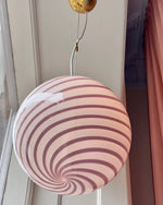 Load image into Gallery viewer, Candy pendant lamp with soft pink and white spiral design, suspended from a brass fixture. The lamp&#39;s swirling pattern adds a playful yet elegant touch to a stylish room with pastel curtains and soft natural light. Its vibrant presence creates a focal point in a modern, minimalist interior that embraces warmth and charm.
