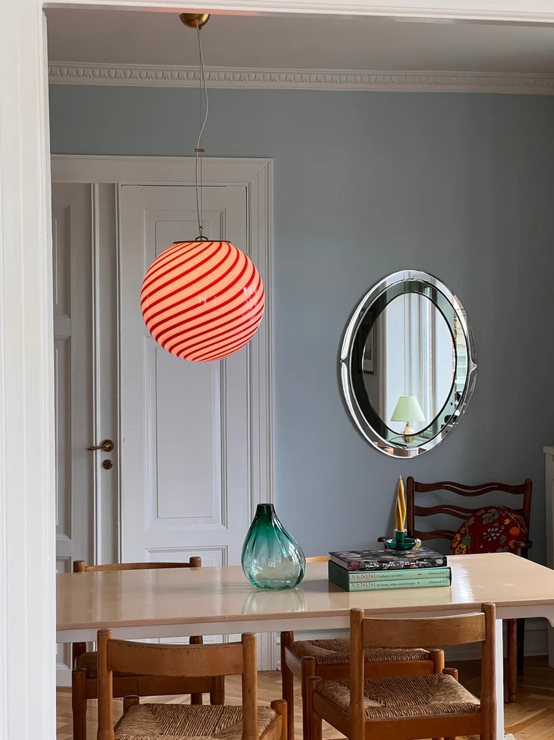 This red and white striped Candy pendant lamp suspended from the ceiling adds a playful touch, complementing a modern and stylish dining space with wooden chairs and a decorative vase.