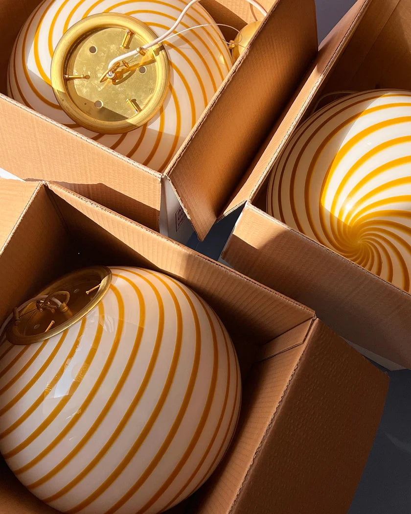 Candy pendant lamp with yellow and white swirl pattern, carefully packed in boxes, showcasing its striking design. The lamp is placed in cardboard packaging, with a hint of brass details peeking through the boxes. The image captures a bright, well-lit room, with sunlight reflecting off the lamp’s glossy surface, adding warmth and charm.